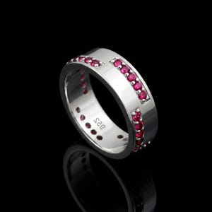 Op de afbeelding: Een zilveren ring met een rij kleine, helderrode edelstenen. De ring heeft een gepolijst, reflecterend oppervlak en staat tegen een zwarte achtergrond. De ring is gemarkeerd met het nummer "925".