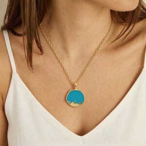 May include: A gold-coloured necklace with a circular pendant. The pendant has a bright turquoise blue enamel centre with a gold-coloured wave design along the bottom edge. The necklace is worn around the neck.