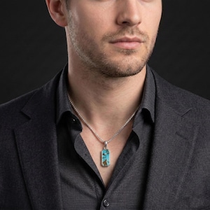 May include: A rectangular pendant featuring a turquoise stone with brown matrix patterns, set in a silver-toned metal frame. The pendant hangs from a silver-toned box chain necklace, resting against a dark gray collared shirt.