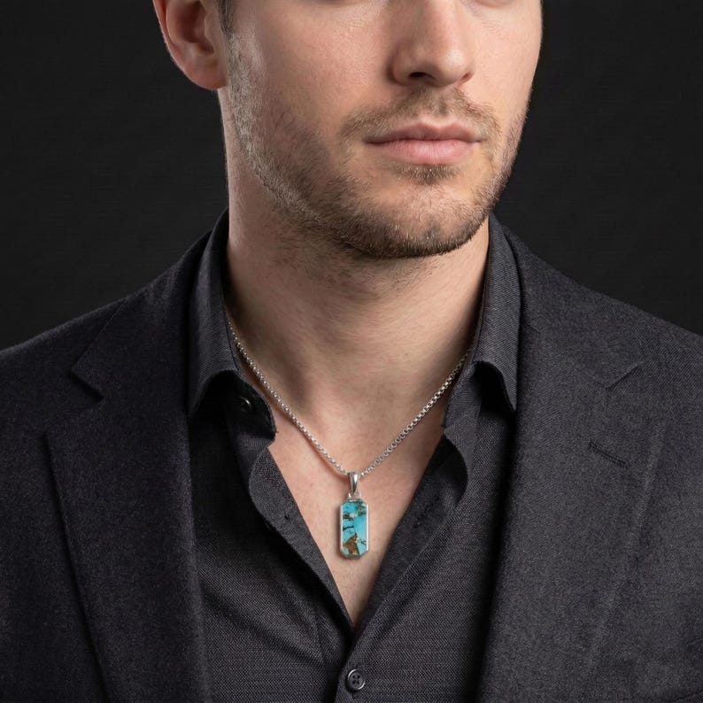 May include: A rectangular pendant featuring a turquoise stone with brown matrix patterns, set in a silver-toned metal frame. The pendant hangs from a silver-toned box chain necklace, resting against a dark gray collared shirt.