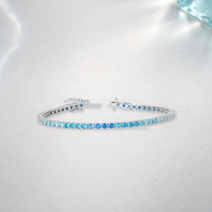 May include: A silver-toned tennis bracelet featuring a continuous row of round-cut gemstones in a gradient of blue hues, ranging from light sky blue to deep azure, secured with a metal box clasp and a safety latch mechanism.