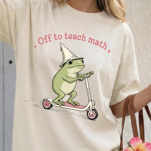 Puede incluir: Camiseta beige con una ilustración caprichosa de una rana verde con un sombrero de mago montada en un patinete rosa y blanco. El texto "Off to teach math" está impreso en rosa encima de la rana.