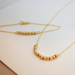 Puede incluir: Un conjunto de collar y pulsera de oro con peque&ntilde;as cuentas redondas. El collar es m&aacute;s largo que la pulsera y las cuentas est&aacute;n dispuestas en forma de curva.