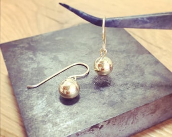 May include: A pair of gold ball drop earrings with a simple design. The earrings are on a gray surface.