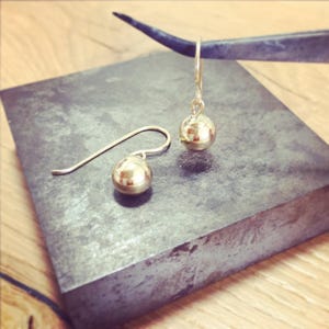 May include: A pair of gold ball drop earrings with a simple design. The earrings are on a gray surface.