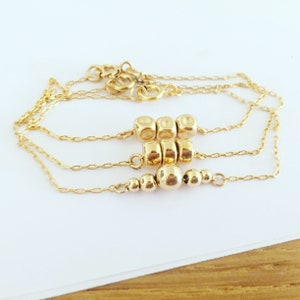 K&ouml;nnte beinhalten: Eine goldene Kettenkette mit drei Reihen goldener Perlen. Die obere Reihe hat drei runde Perlen, die mittlere Reihe vier runde Perlen und die untere Reihe f&uuml;nf runde Perlen.