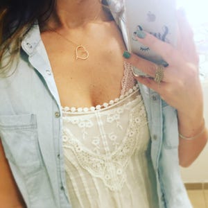 May include: A woman wearing a light blue denim shirt over a white lace top with a delicate gold heart necklace.