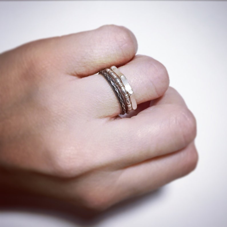 May include: Three silver rings stacked on a finger. The top ring is a thin, flat band. The middle ring is a textured band with small beads. The bottom ring is a braided band.