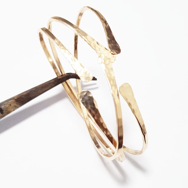 Puede incluir: Tres pulseras de metal martillado de color dorado, apiladas y entrelazadas. Las pulseras son abiertas y tienen una forma ligeramente curvada.