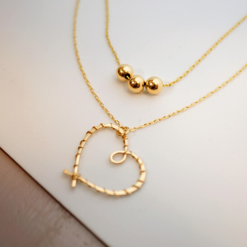 May include: A gold necklace with a heart pendant and three round beads. The necklace is on a white surface.