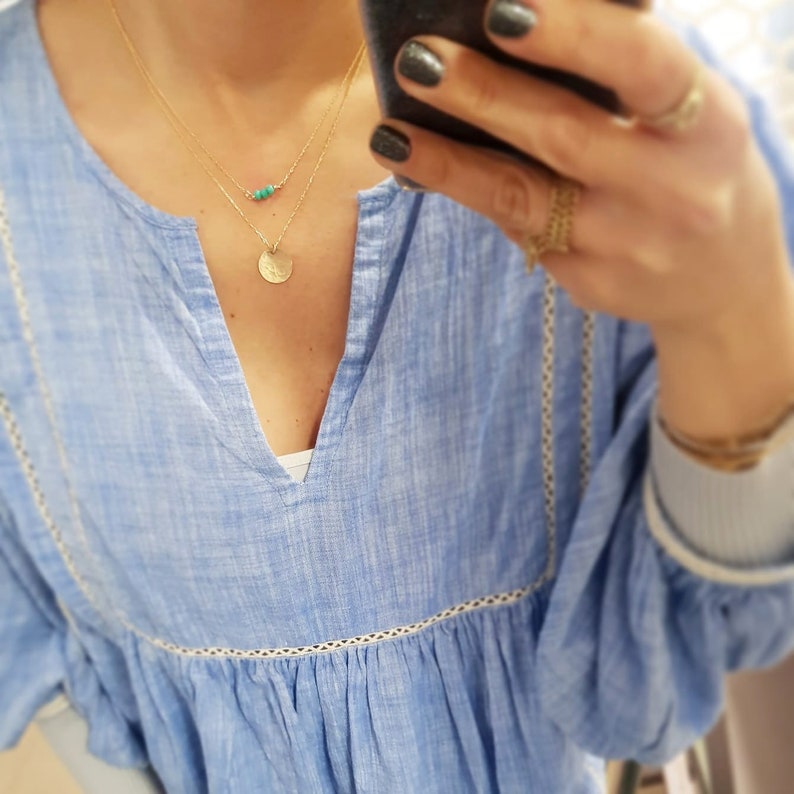 May include: A light blue, loose-fitting, cotton blouse with a V-neckline and white lace trim. The blouse features a gold chain necklace with a small turquoise stone pendant and a larger circular pendant.
