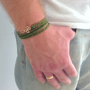 May include: A green braided bracelet with a gold charm. The bracelet is worn on a person's wrist.