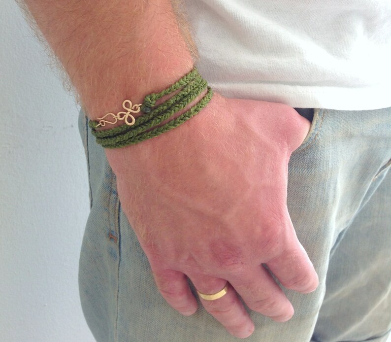 May include: A green braided bracelet with a gold charm. The bracelet is worn on a person's wrist.