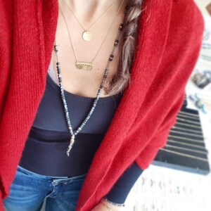 May include: A woman wearing a red cardigan over a black tank top and blue jeans. She is wearing a layered gold necklace with a round pendant and a fringe pendant. She is also wearing a long black beaded necklace.