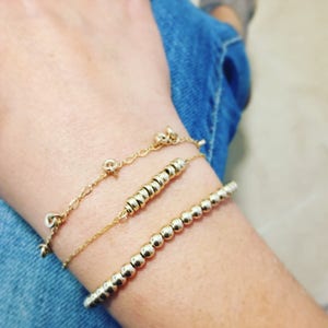 May include: Three gold bracelets on a wrist. The top bracelet is a delicate chain with small charms. The middle bracelet is a stack of small gold rings. The bottom bracelet is a string of round gold beads.