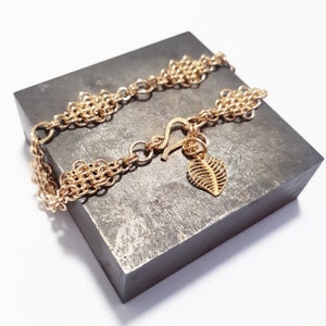 May include: A gold chain bracelet with a leaf-shaped charm. The bracelet is made of multiple strands of chain and has a lobster clasp.