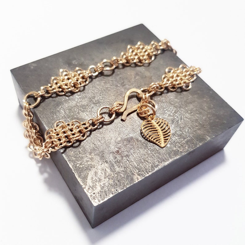 May include: A gold chain bracelet with a leaf-shaped charm. The bracelet is made of multiple strands of chain and has a lobster clasp.