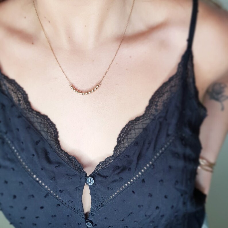 May include: A gold necklace with a delicate chain and a row of small, round beads. The necklace is worn by a person with a black top.