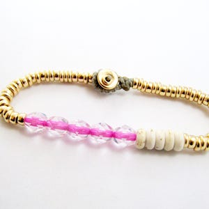 May include: Gold beaded bracelet with a pink and clear crystal section, and a section of white beads. The bracelet is secured with a gold clasp and a gray cord. Jewelry.