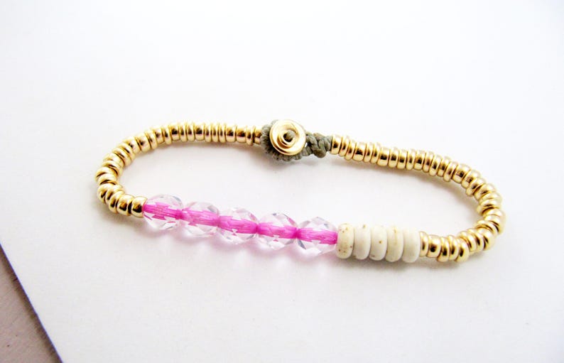 May include: Gold beaded bracelet with a pink and clear crystal section, and a section of white beads. The bracelet is secured with a gold clasp and a gray cord. Jewelry.