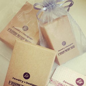 May include: Two brown cardboard boxes with the text "Jewelry + Designer" and "Jewelry + Designer" printed on them. The boxes are wrapped in a sheer white fabric with a brown ribbon.