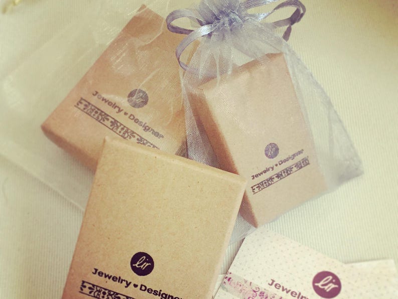 May include: Two brown cardboard boxes with the text "Jewelry + Designer" and "Jewelry + Designer" printed on them. The boxes are wrapped in a sheer white fabric with a brown ribbon.