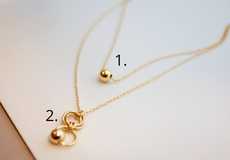 May include: Two gold necklaces with different pendants. The first necklace has a single round pendant. The second necklace has a pendant with three interlocking circles and a round bead.