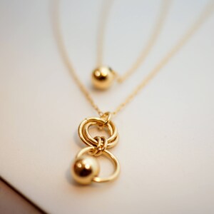 May include: A gold necklace with a layered chain and a pendant featuring two interlocking circles and a small ball charm.