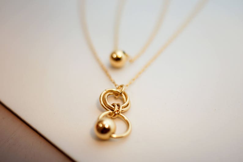 May include: A gold necklace with a layered chain and a pendant featuring two interlocking circles and a small ball charm.
