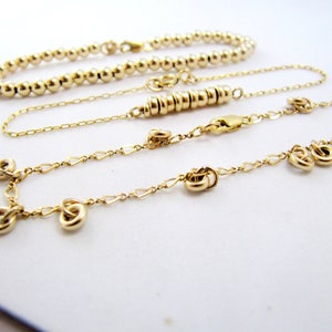 May include: Three gold necklaces, one with a beaded chain, one with a delicate chain, and one with a chain with small knot-shaped charms.