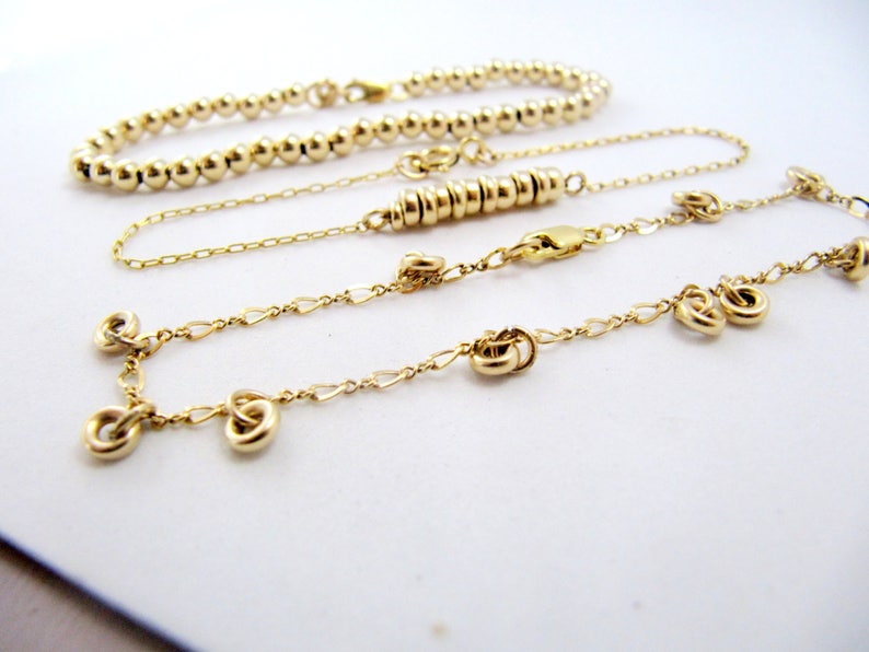 May include: Three gold necklaces, one with a beaded chain, one with a delicate chain, and one with a chain with small knot-shaped charms.