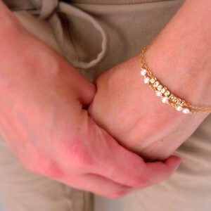 Puede incluir: Un brazalete de cadena dorada con peque&ntilde;as cuentas blancas y un dise&ntilde;o delicado. El brazalete se lleva en la mu&ntilde;eca.