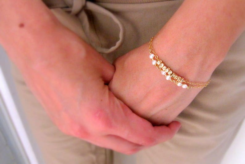 Puede incluir: Un brazalete de cadena dorada con peque&ntilde;as cuentas blancas y un dise&ntilde;o delicado. El brazalete se lleva en la mu&ntilde;eca.