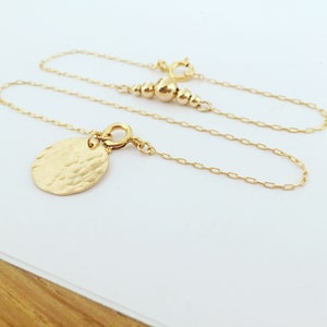 Puede incluir: Un delicado collar de oro con un colgante ovalado martillado. El collar presenta una fina cadena y una serie de peque&ntilde;as cuentas doradas cerca del cierre. El colgante tiene una superficie texturizada, a&ntilde;adiendo un toque &uacute;nico a la joya.