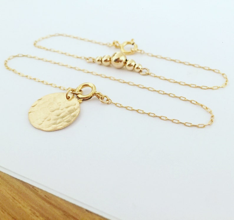 Puede incluir: Un delicado collar de oro con un colgante ovalado martillado. El collar presenta una fina cadena y una serie de peque&ntilde;as cuentas doradas cerca del cierre. El colgante tiene una superficie texturizada, a&ntilde;adiendo un toque &uacute;nico a la joya.