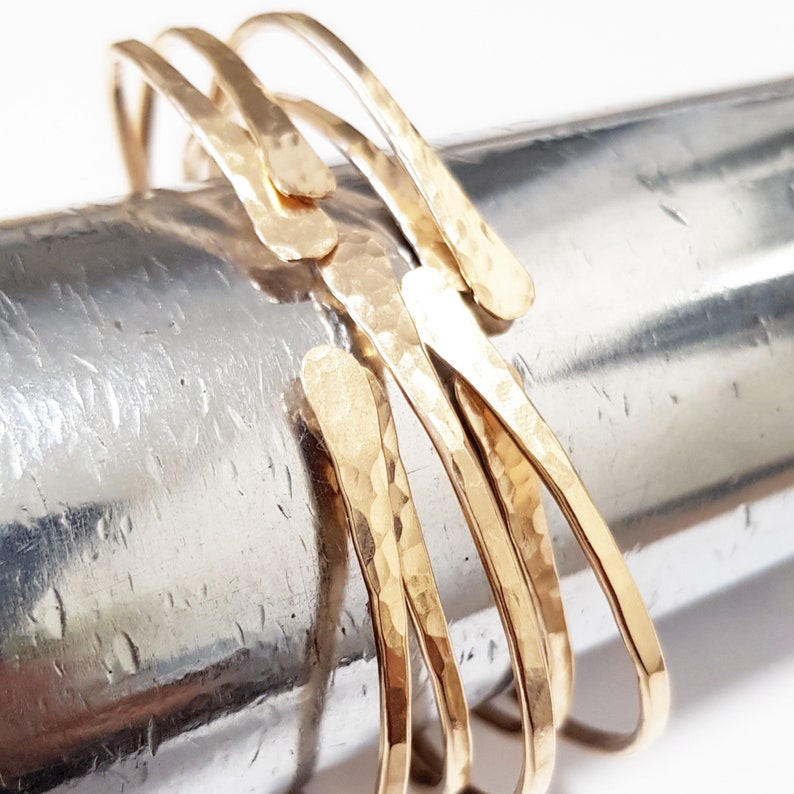 Puede incluir: Un conjunto de cuatro pulseras de metal martillado de color dorado. Las pulseras est&aacute;n apiladas una encima de la otra y tienen una forma ligeramente irregular.