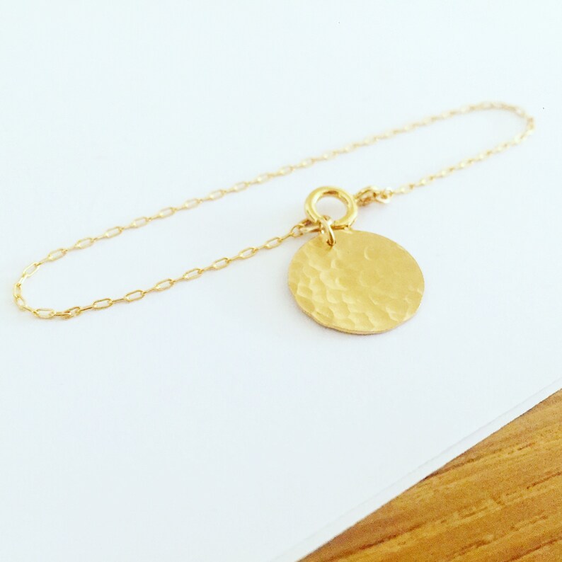 Puede incluir: Una delicada pulsera dorada con un colgante circular con textura martillada. El colgante est&aacute; unido a una fina cadena con un peque&ntilde;o anillo. La pulsera se muestra sobre una superficie blanca.