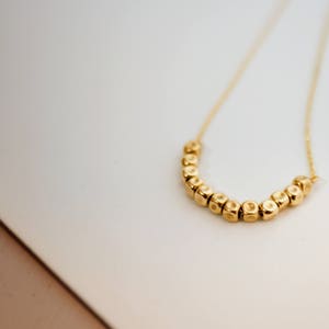 Puede incluir: Un collar de oro con una cadena y una fila de peque&ntilde;as cuentas doradas texturizadas en forma de U.