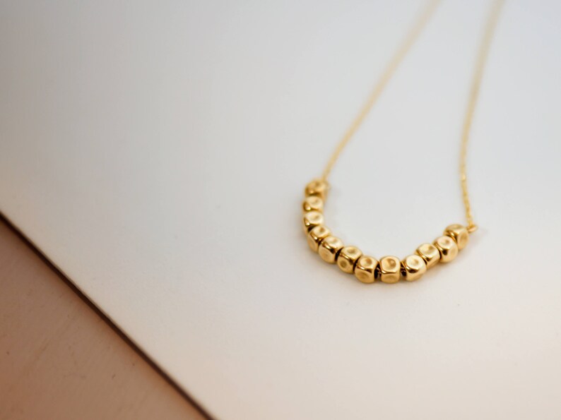 Puede incluir: Un collar de oro con una cadena y una fila de peque&ntilde;as cuentas doradas texturizadas en forma de U.