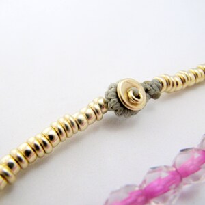 May include: Close-up of a gold beaded bracelet with a gray knot and a gold clasp. The bracelet also features a section of pink and clear faceted beads. The jewelry is set against a white background.