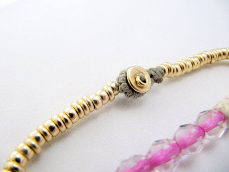 May include: Close-up of a gold beaded bracelet with a gray knot and a gold clasp. The bracelet also features a section of pink and clear faceted beads. The jewelry is set against a white background.