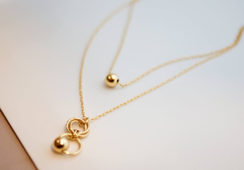 May include: A gold necklace with two chains. The longer chain has a single gold ball pendant. The shorter chain has a pendant with three interlocking gold circles and a gold ball.