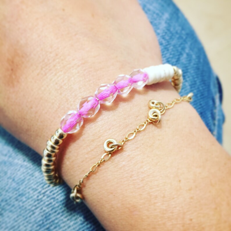 May include: Two bracelets are shown. One is a gold-toned metal bracelet with a white woven section and pink beads. The other is a delicate gold chain bracelet. Both bracelets are on a wrist, with a denim background.