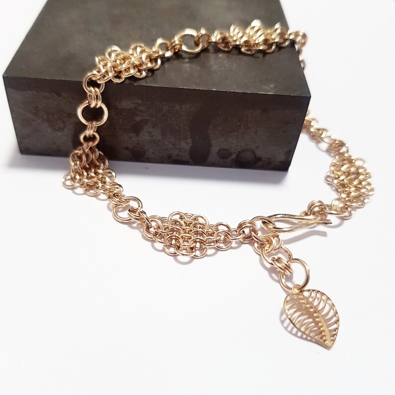 May include: A gold chain bracelet with a delicate leaf charm. The chain is made of multiple strands of small, interconnected rings.