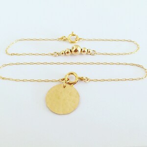 Puede incluir: Pulsera de cadena dorada con un dije de disco martillado. La pulsera presenta un dise&ntilde;o de cadena delicado y un peque&ntilde;o dije redondo. Tambi&eacute;n se ve una segunda cadena dorada con peque&ntilde;as cuentas doradas.