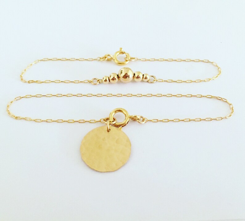 Puede incluir: Pulsera de cadena dorada con un dije de disco martillado. La pulsera presenta un dise&ntilde;o de cadena delicado y un peque&ntilde;o dije redondo. Tambi&eacute;n se ve una segunda cadena dorada con peque&ntilde;as cuentas doradas.