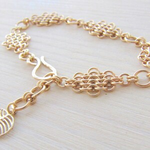 May include: Gold chain bracelet with a leaf charm. The bracelet is made of interwoven metal rings and has a lobster claw clasp.