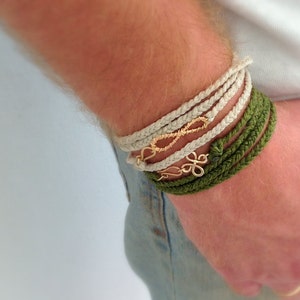May include: A person wearing a stack of bracelets on their wrist. The bracelets are made of braided rope in white and green. The bracelets are adorned with gold infinity and flower charms.