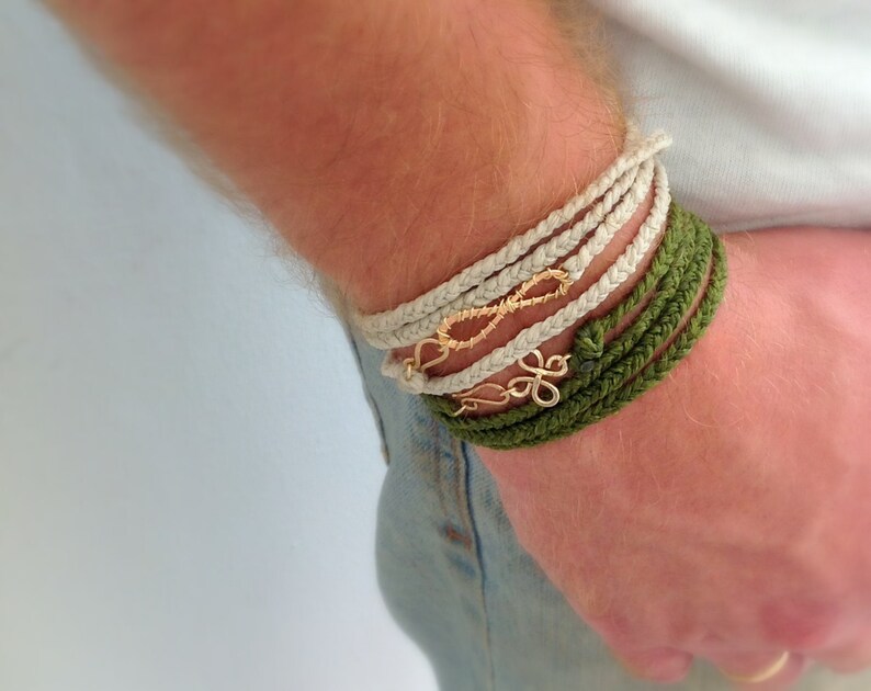 May include: A person wearing a stack of bracelets on their wrist. The bracelets are made of braided rope in white and green. The bracelets are adorned with gold infinity and flower charms.