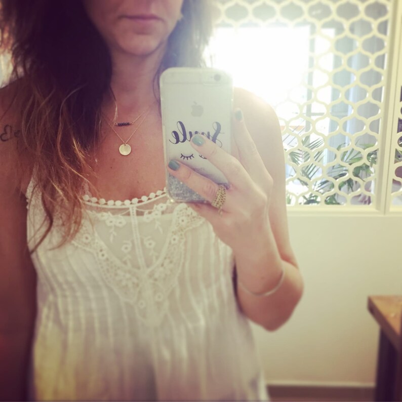 May include: A woman wearing a white lace top with a gold necklace and a gold ring. She is holding a phone with a glitter case that says "Smile" with a green and black eye design.